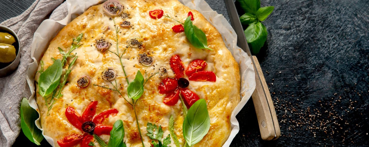 Focaccia mit Tomaten, Basilikum und weiteren Belägen auf Backpapier