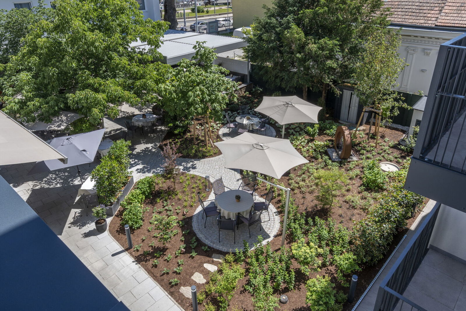 Begrünter Innenhof des Restaurants Der Stasta in Wien-Liesing mit Bäumen, bepflanzten Beeten, Sonnenschirmen und Gastgartentischen.